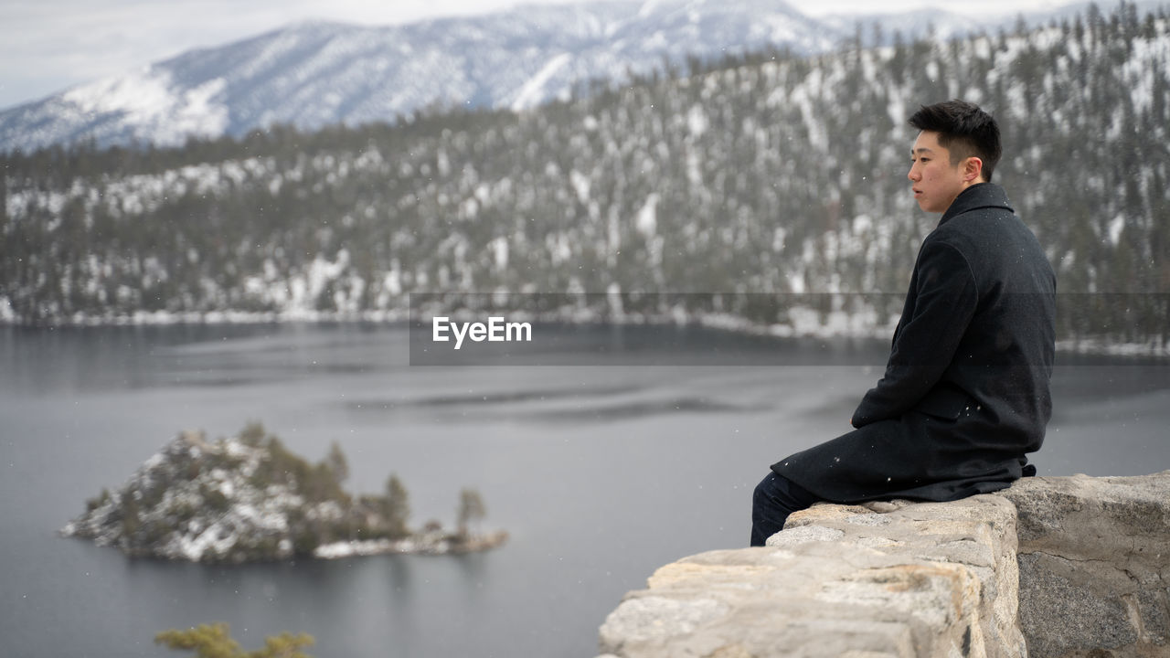 Young man looking away while sitting on retaining wall over lake