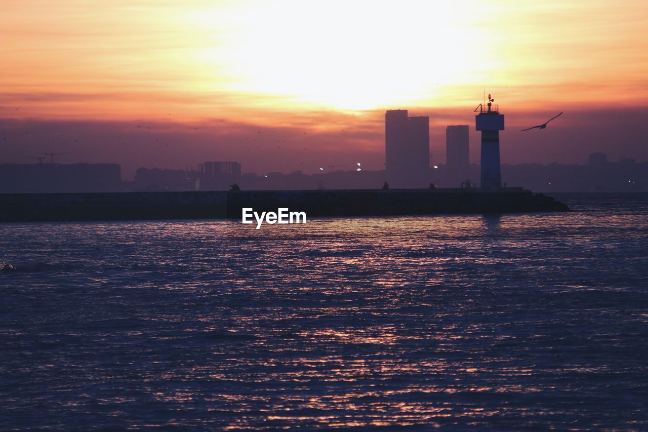 Silhouette buildings by sea against sky during sunset