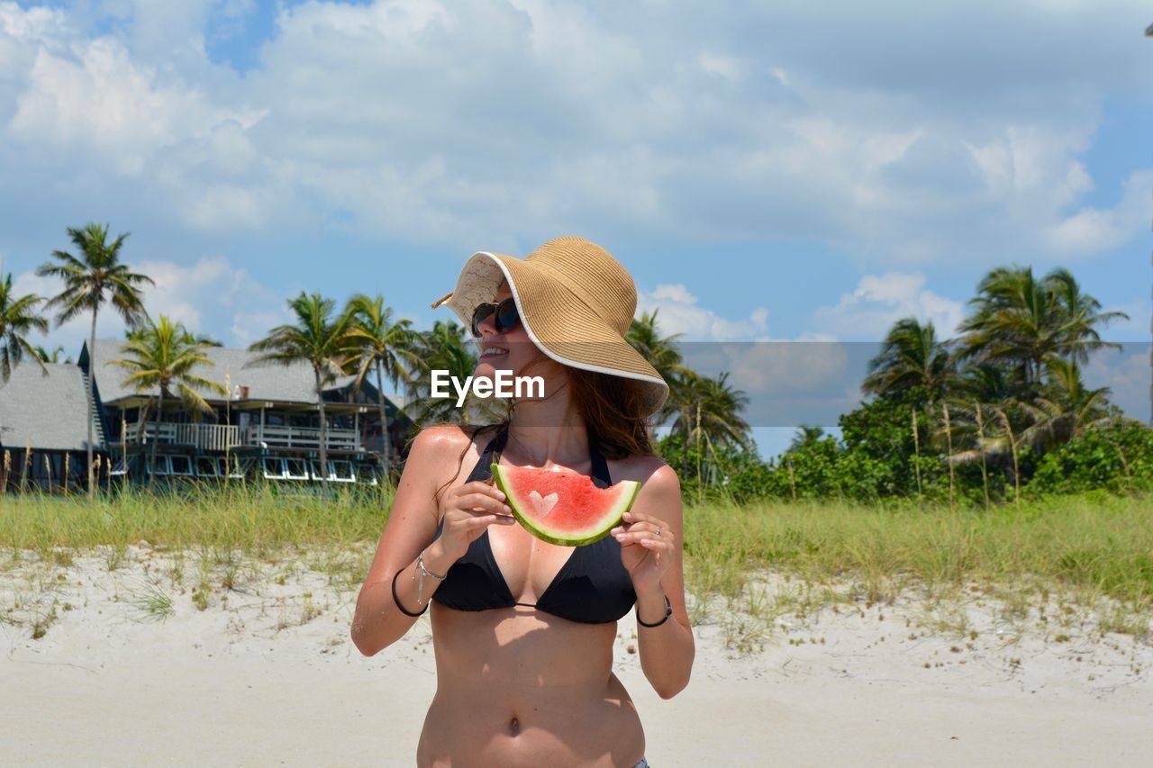 Young woman wearing bikini holding watermelon while standing at beach against cloudy sky