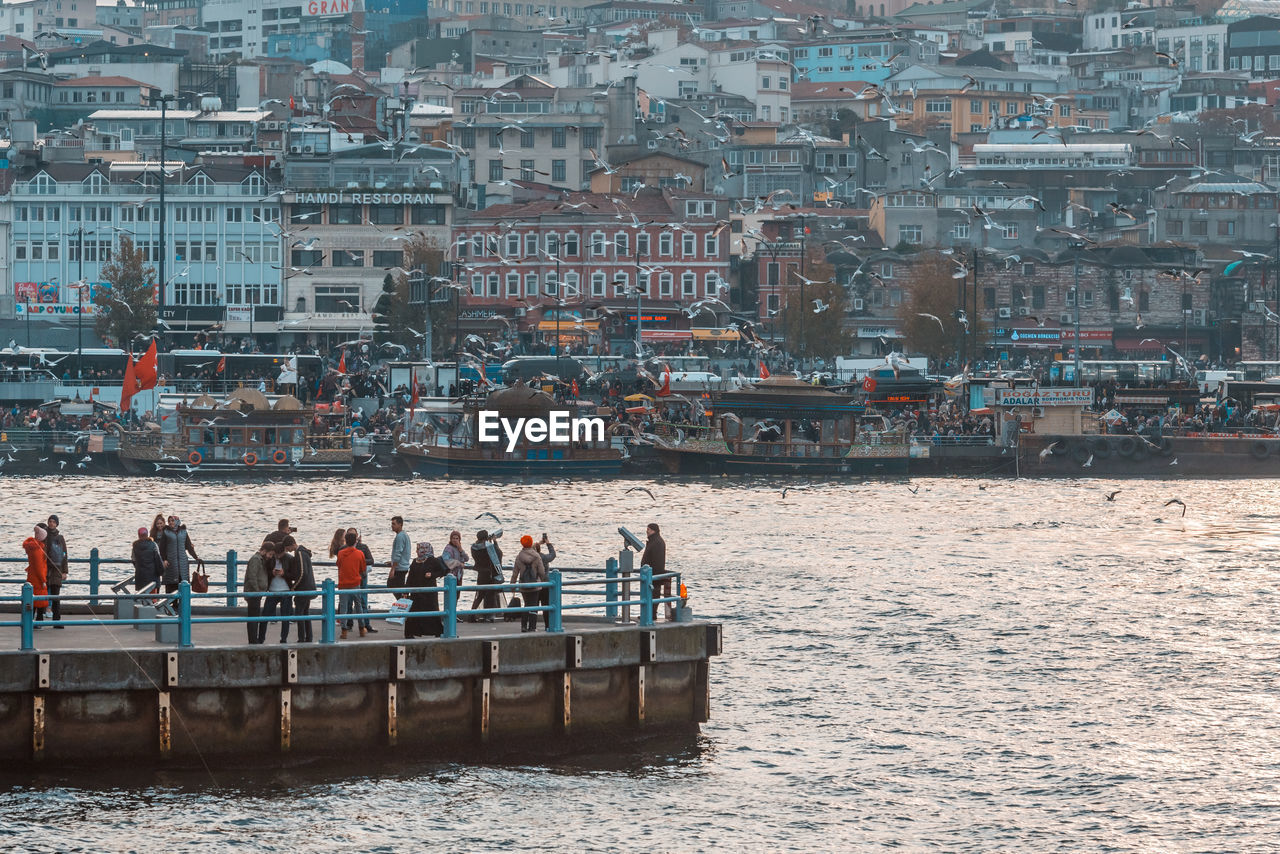 GROUP OF PEOPLE IN CITY BY SEA AGAINST BUILDINGS IN BACKGROUND