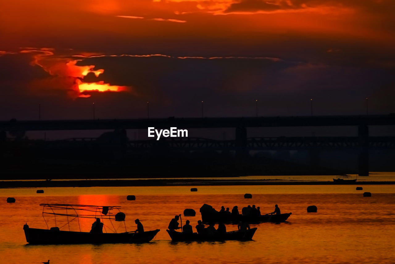 Silhouette people sitting on rowboats in river against sky during sunset