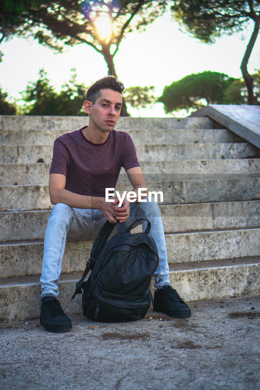 Portrait of young man holding backpack while sitting on staircase