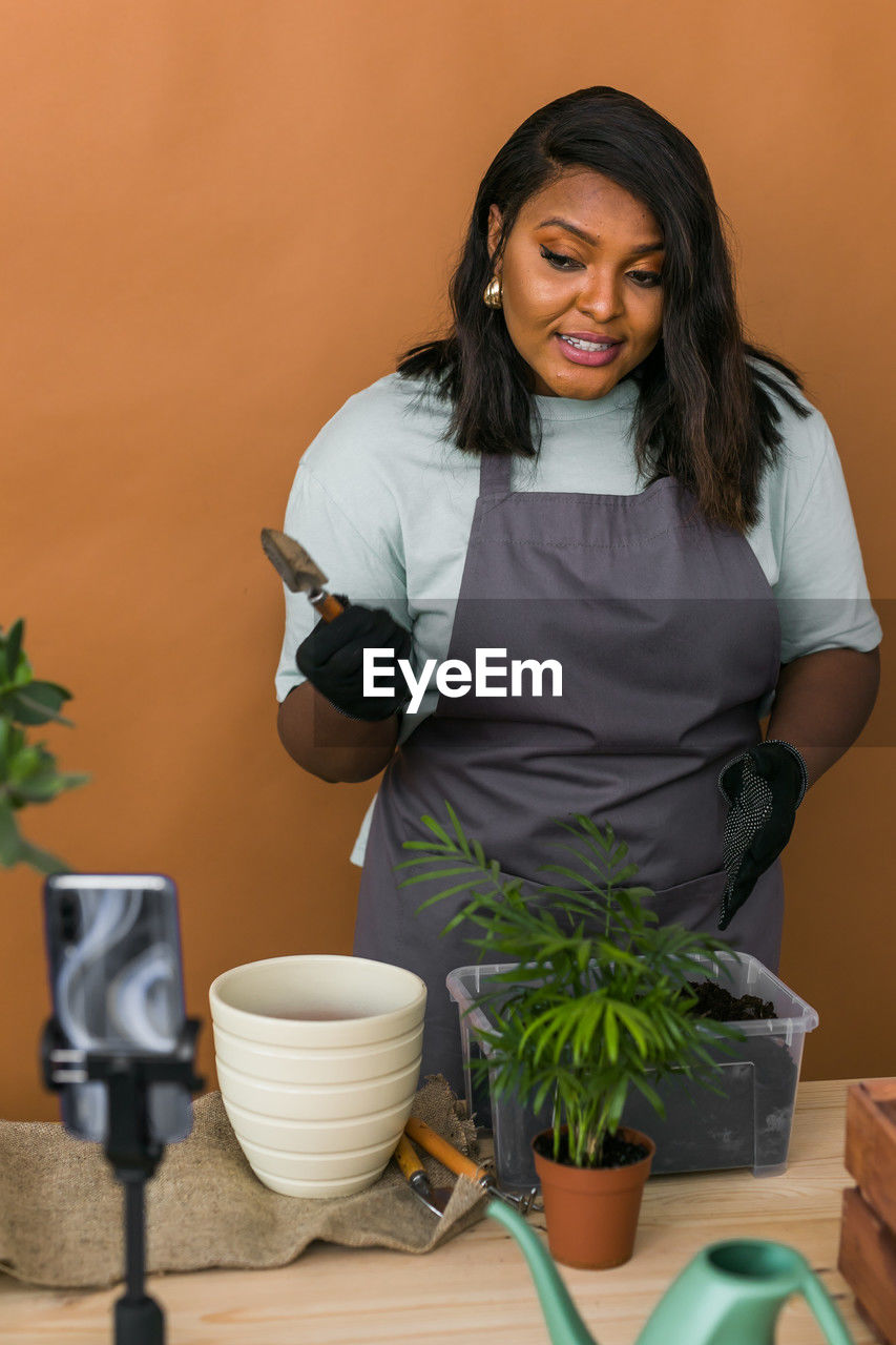 women, adult, one person, potted plant, smiling, indoors, female, lifestyles, plant, standing, happiness, portrait, growth, food and drink, front view, holding, looking at camera, occupation, domestic life, long hair, cheerful, emotion, working, business, young adult, black hair, nature, houseplant, flowerpot, food, hairstyle, person, apron, wellbeing, three quarter length, business finance and industry, clothing, gardening, small business, protective glove