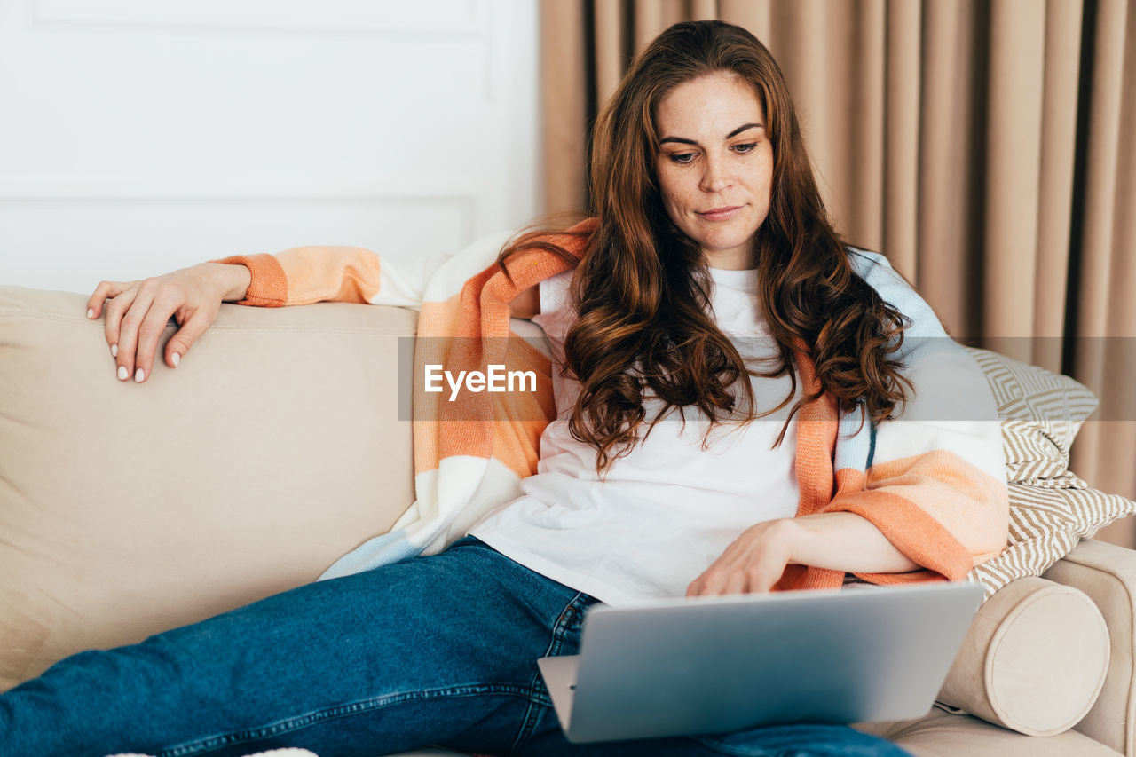 Young concentrated business woman lying on sofa at home uses laptop to watch movie.