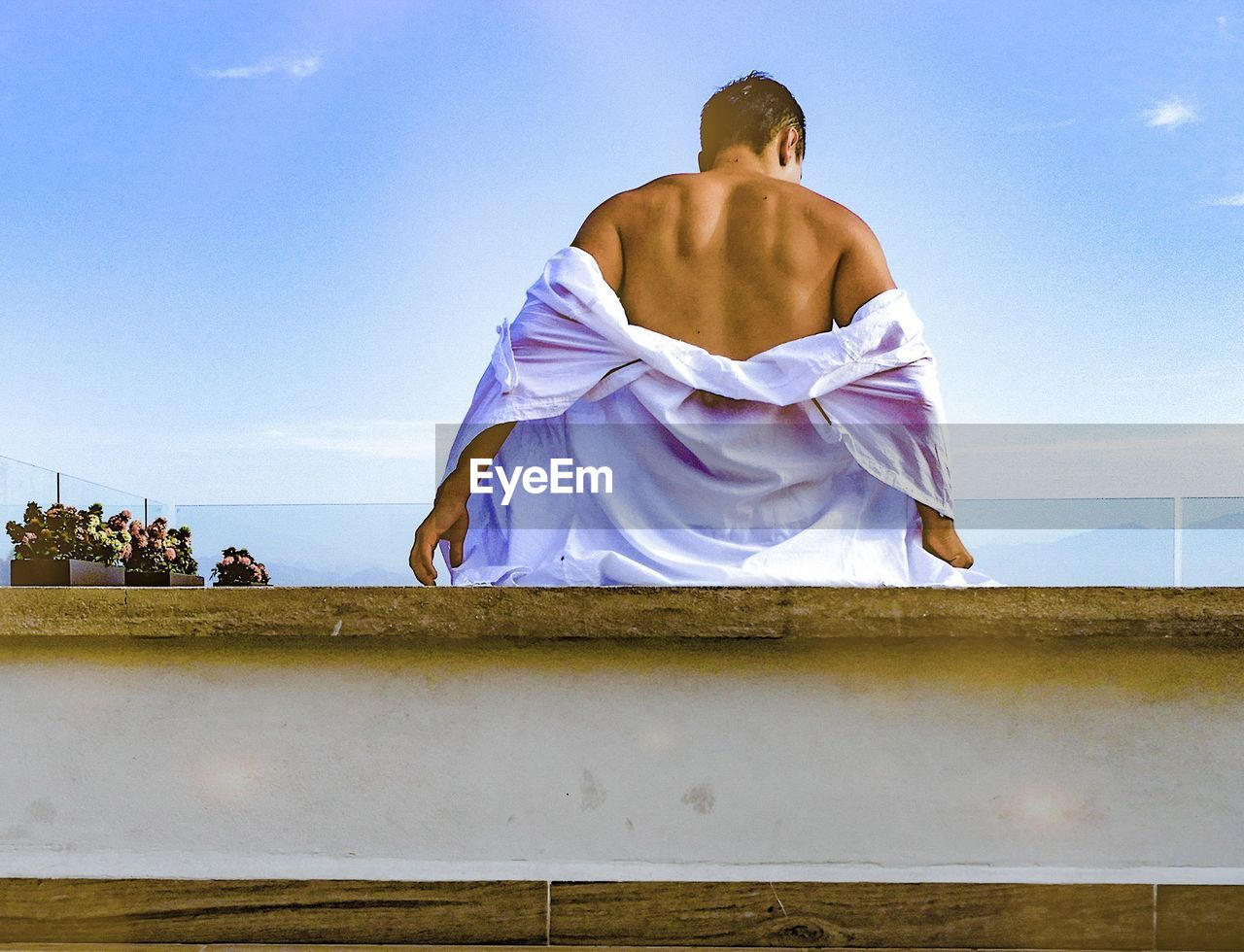 REAR VIEW OF SHIRTLESS MAN SITTING AGAINST SKY