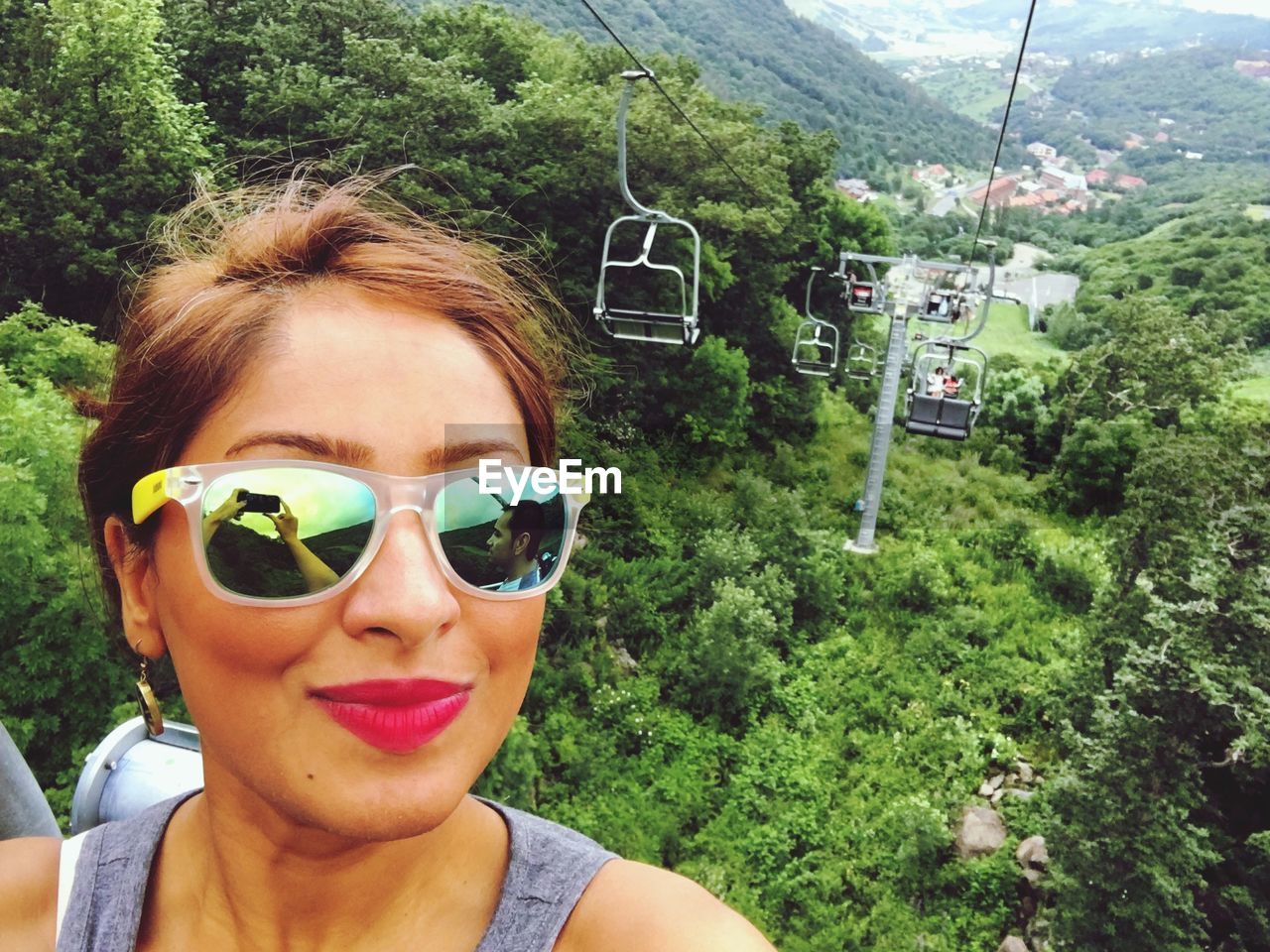 Portrait of young woman wearing sunglasses with reflection against ski lift over trees