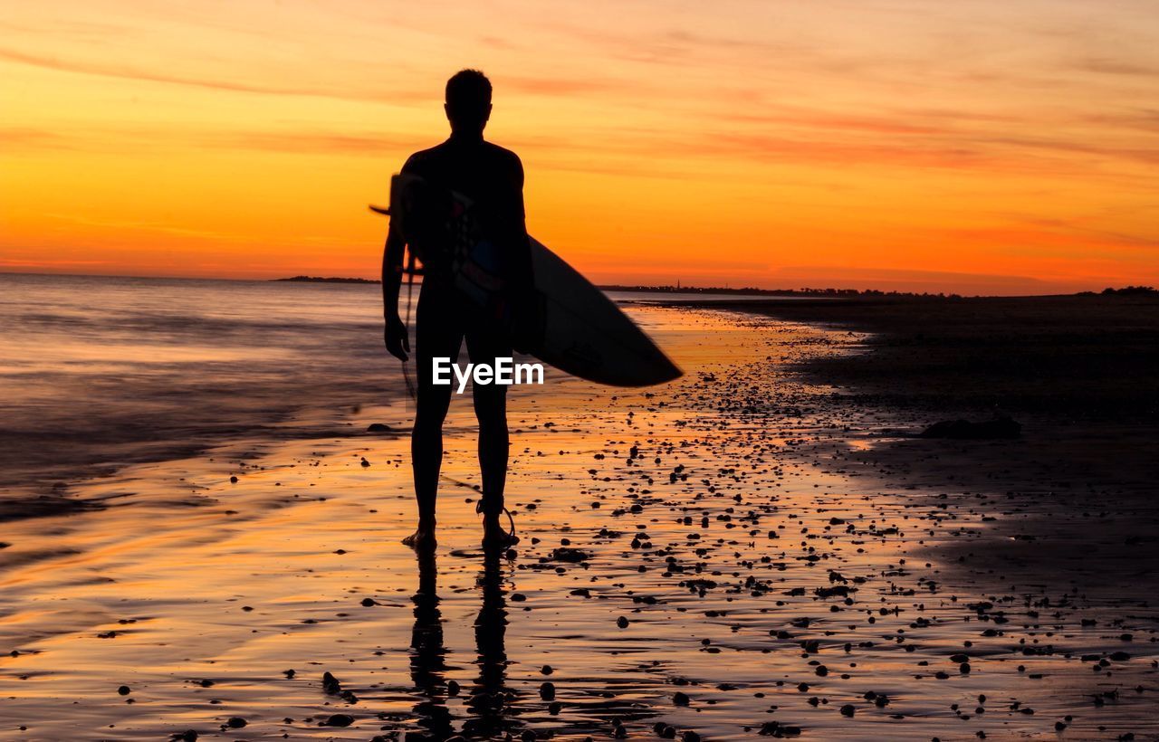 Rear view of silhouette man with surfboard standing on shore at beach during sunset