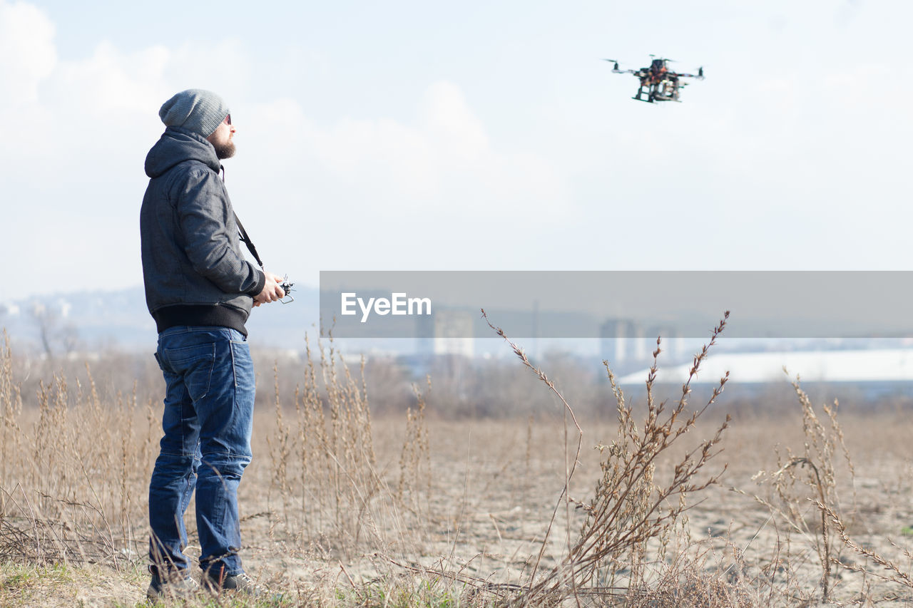 Side view of mid adult man flying drone while standing on field against sky