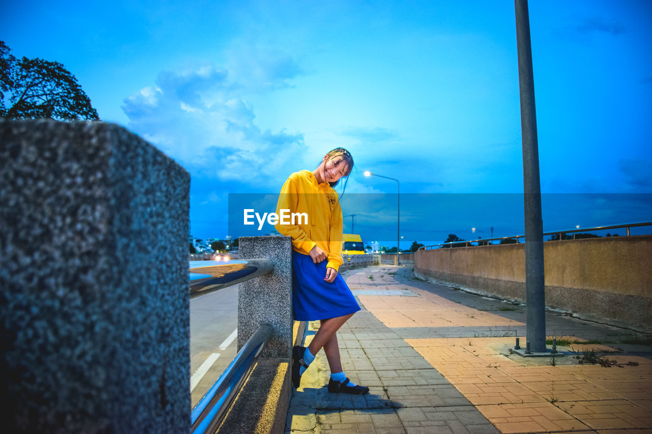 Portrait of smiling young woman standing on footpath against blue sky