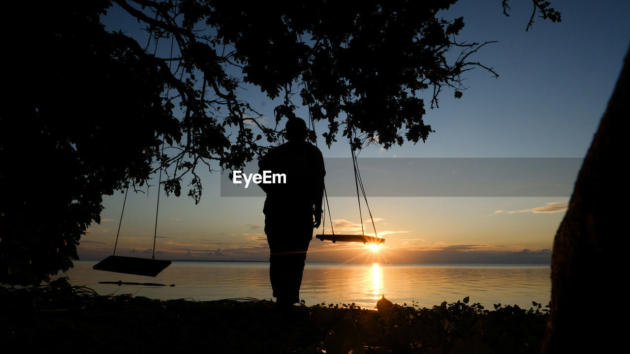 water, silhouette, sky, morning, sea, nature, beach, sunlight, tree, beauty in nature, dusk, land, backlighting, one person, reflection, plant, tranquility, scenics - nature, horizon, adult, tranquil scene, sunrise, sun, men, lifestyles, darkness, light, leisure activity, standing, outdoors, rear view, horizon over water, vacation, idyllic, holiday, cloud, trip, person, full length