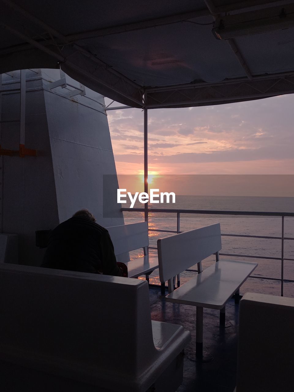 REAR VIEW OF MAN SITTING ON BOAT AT SEA SHORE AGAINST SKY