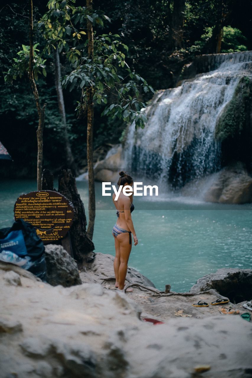 FULL LENGTH OF WOMAN STANDING ON ROCKS BY WATERFALL