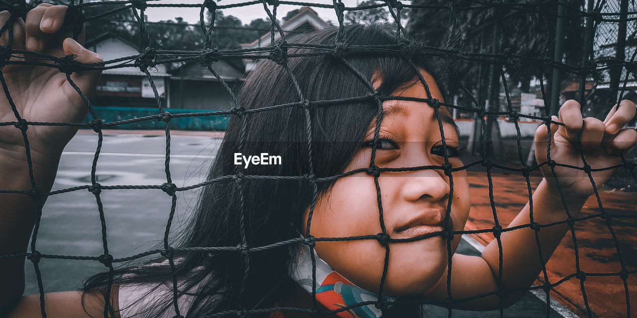 CLOSE-UP PORTRAIT OF YOUNG WOMAN STANDING BY FENCE AGAINST METAL STRUCTURE