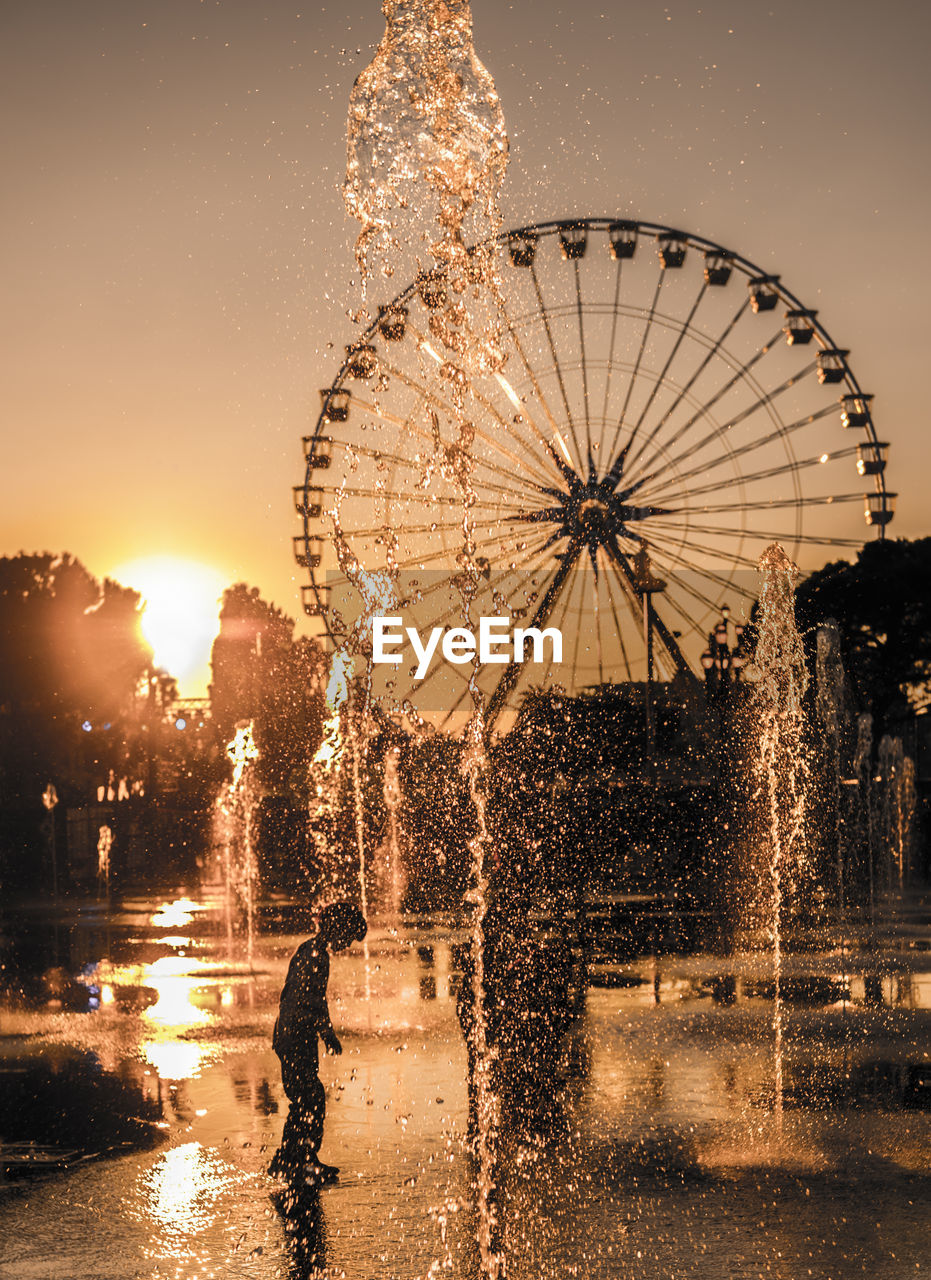 Child standing at fountain against ferris wheel during sunset