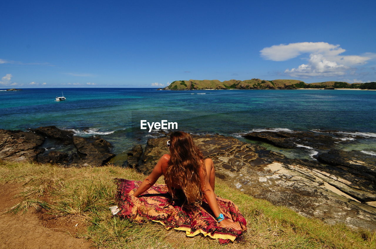 REAR VIEW OF WOMAN SITTING AT BEACH AGAINST CLEAR BLUE SKY