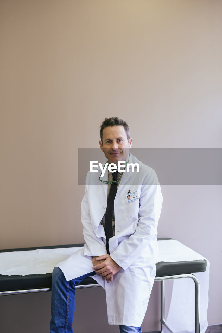 Portrait of smiling doctor sitting on bed against wall