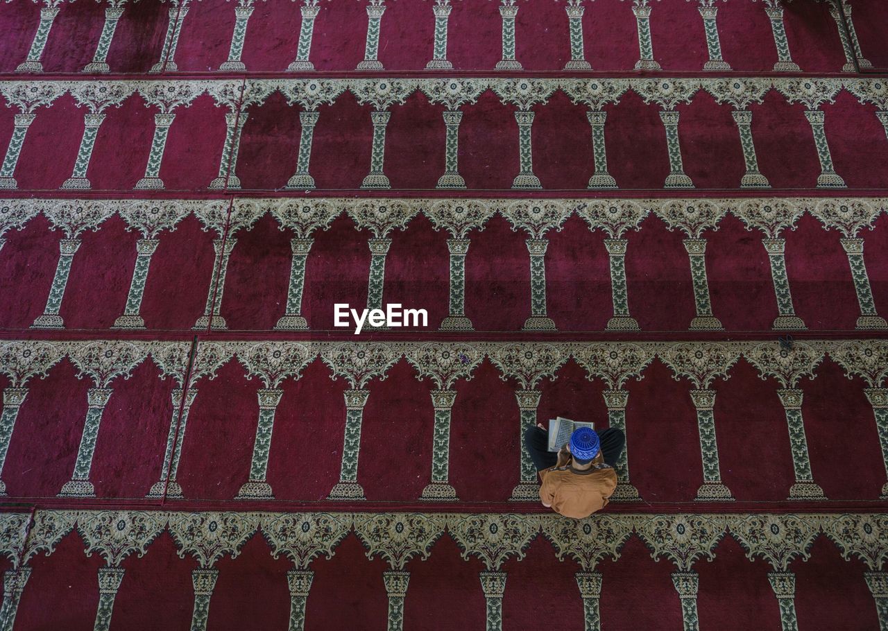 High angle view of man reading quran while sitting on carpet in mosque