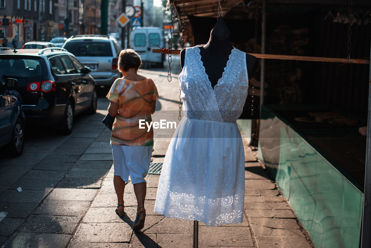 Rear view full length of woman walking by mannequin on sidewalk in city