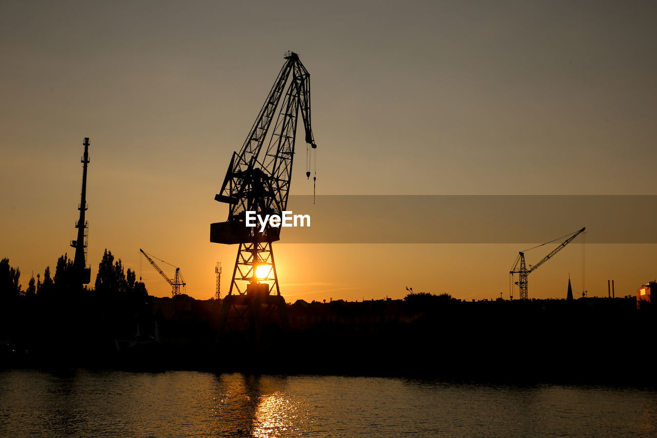 Silhouette cranes by river against sky during sunset