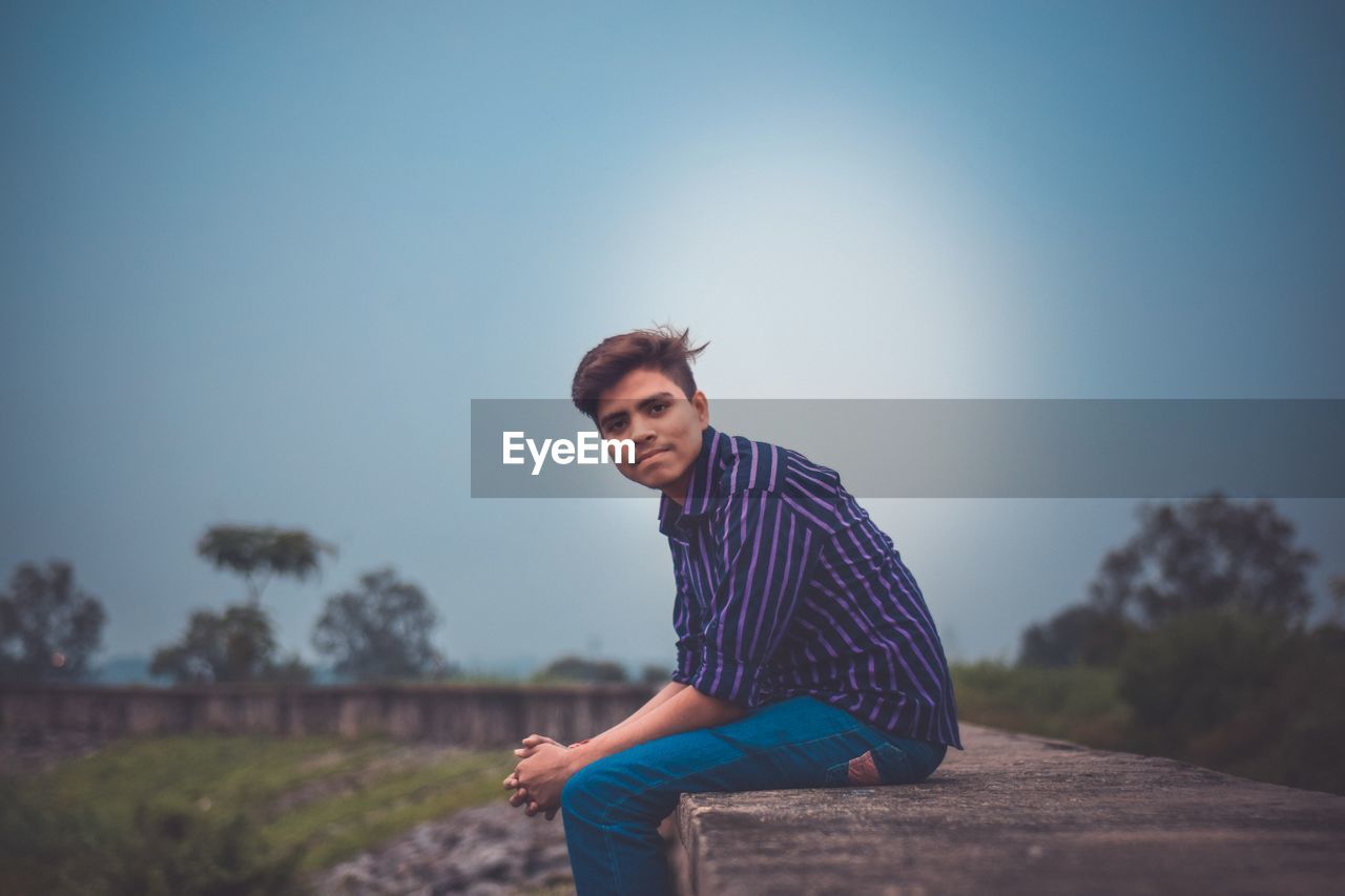 Portrait of young man sitting on retaining wall against sky