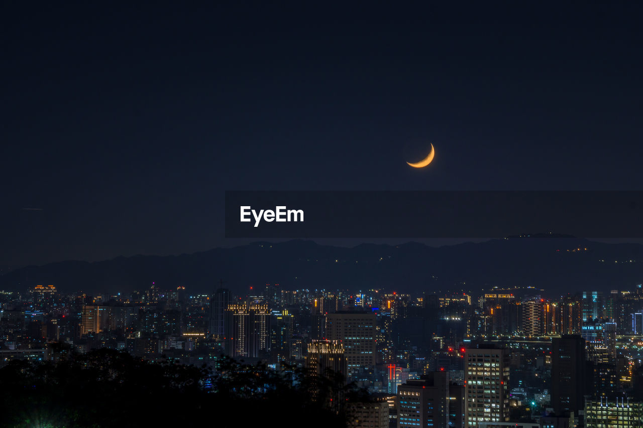 Illuminated city against sky at night with a crescent moon