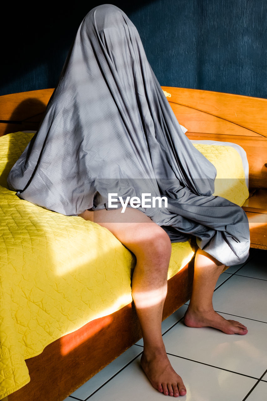 One lonely person sitting on bed while covering his face with a bed sheet