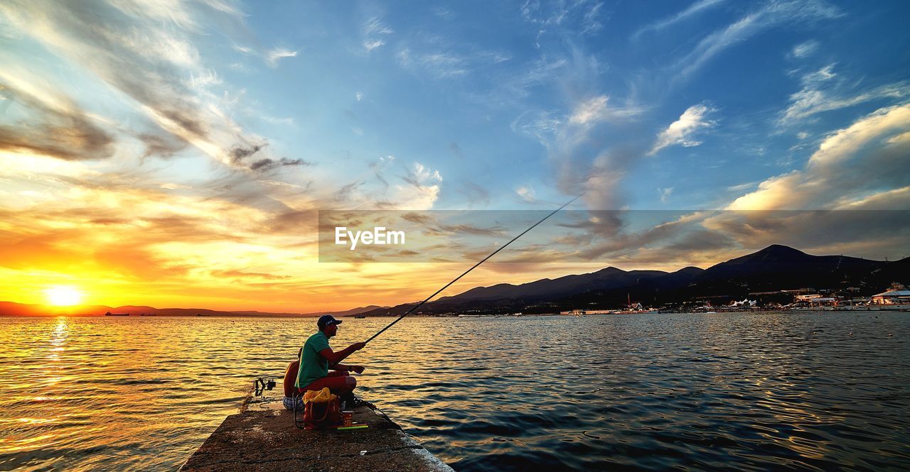 Man fishing in sea against sky during sunset