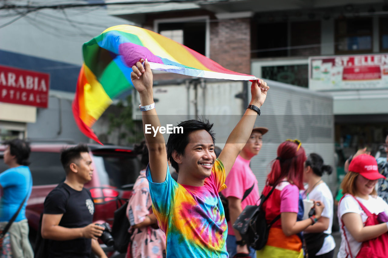 Smiling man holding rainbow flag while standing in city