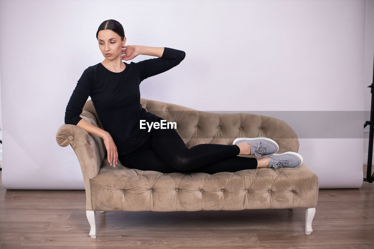 sitting, adult, indoors, one person, full length, limb, furniture, lifestyles, women, couch, human leg, relaxation, person, portrait, young adult, female, photo shoot, clothing, living room, studio couch, looking at camera, smiling, black, wellbeing, happiness, wood, physical fitness, footwear