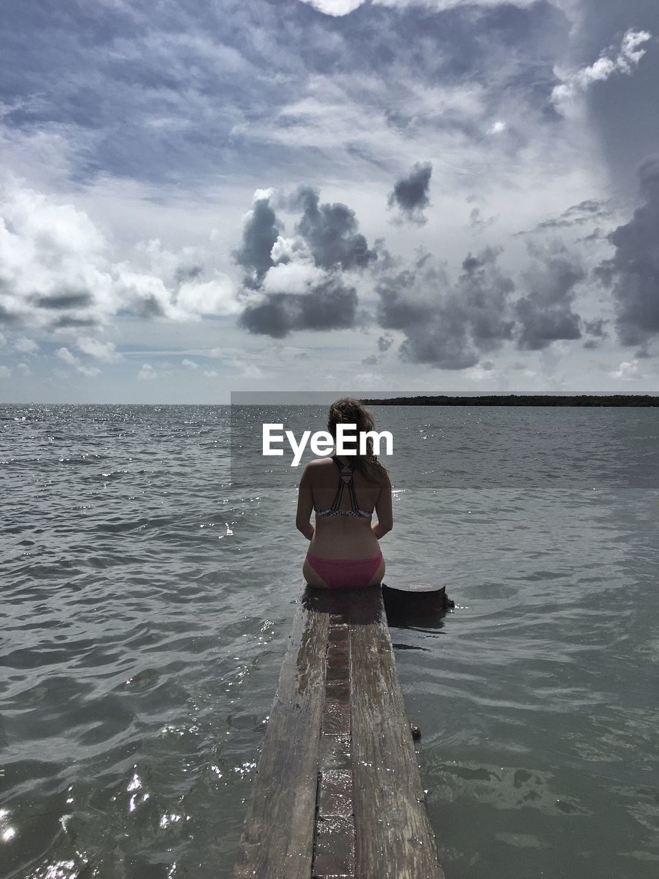 Rear view of young woman sitting on jetty over sea against cloudy sky