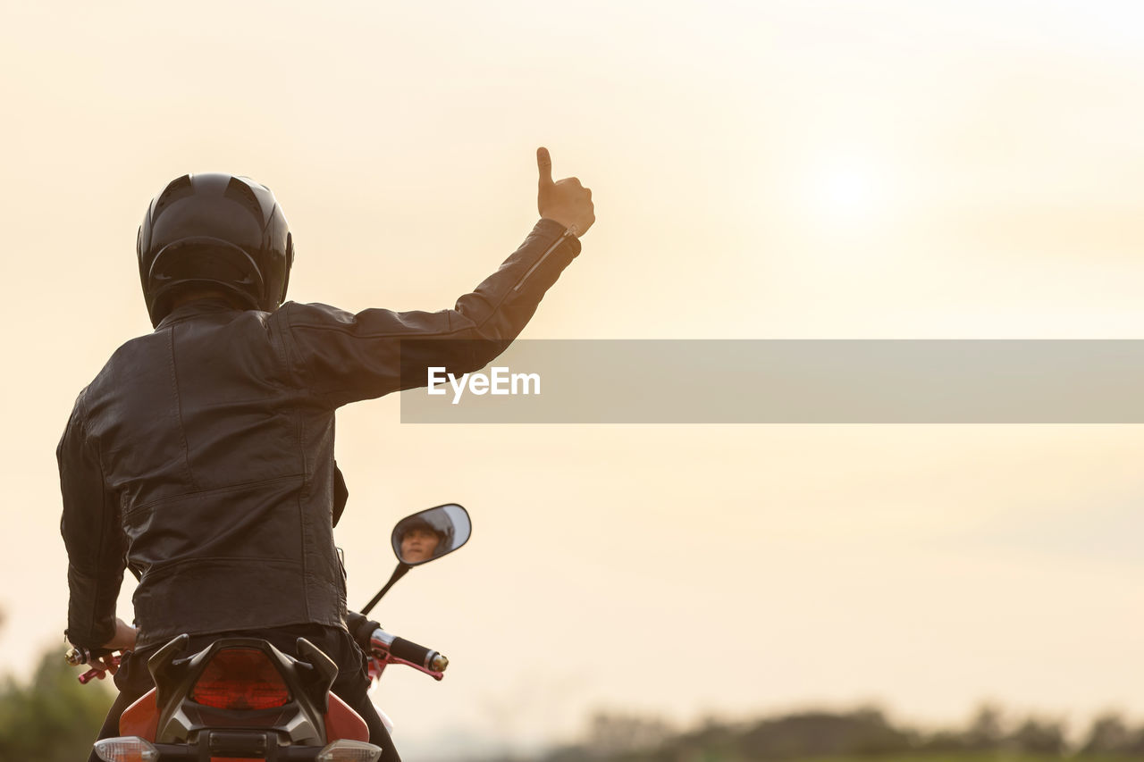 Rear view of man showing thumbs up sign while sitting on motorcycle against clear sky during sunset