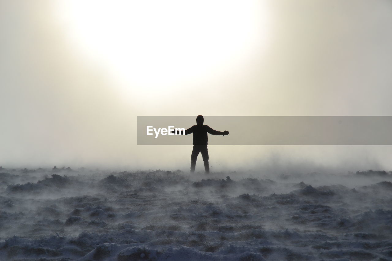 Silhouette man with arms outstretched standing in fog against sky