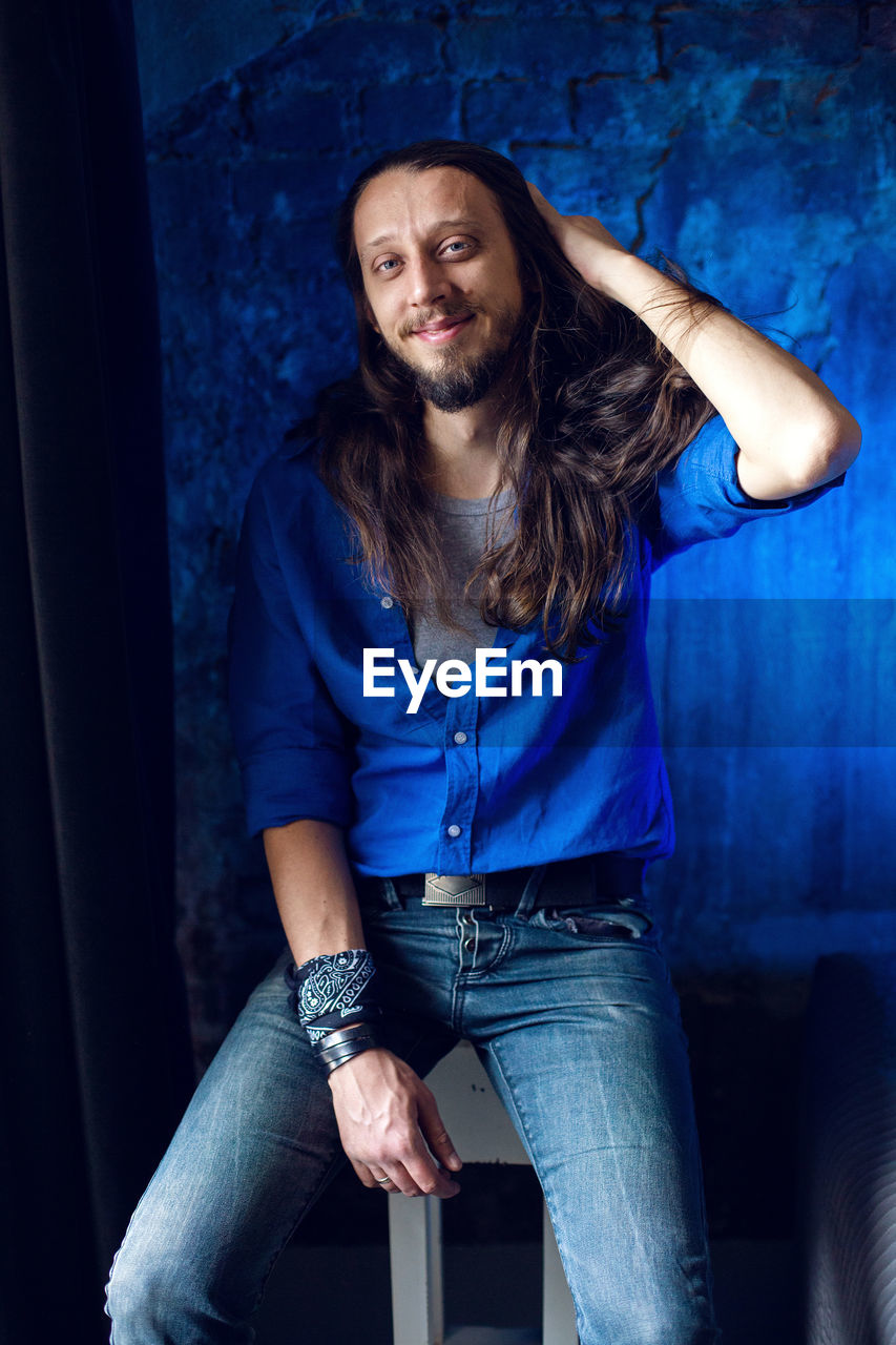 Man with long hair and beard sits at home on a chair by the window in a blue shirt and jeans against