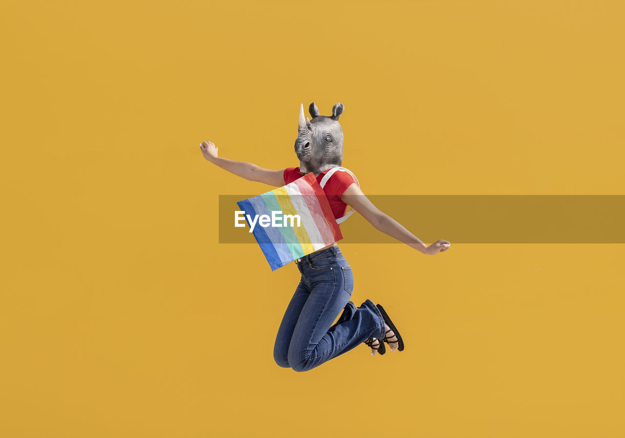 Woman wearing rhinoceros mask jumping with multi colored bag in front of yellow wall