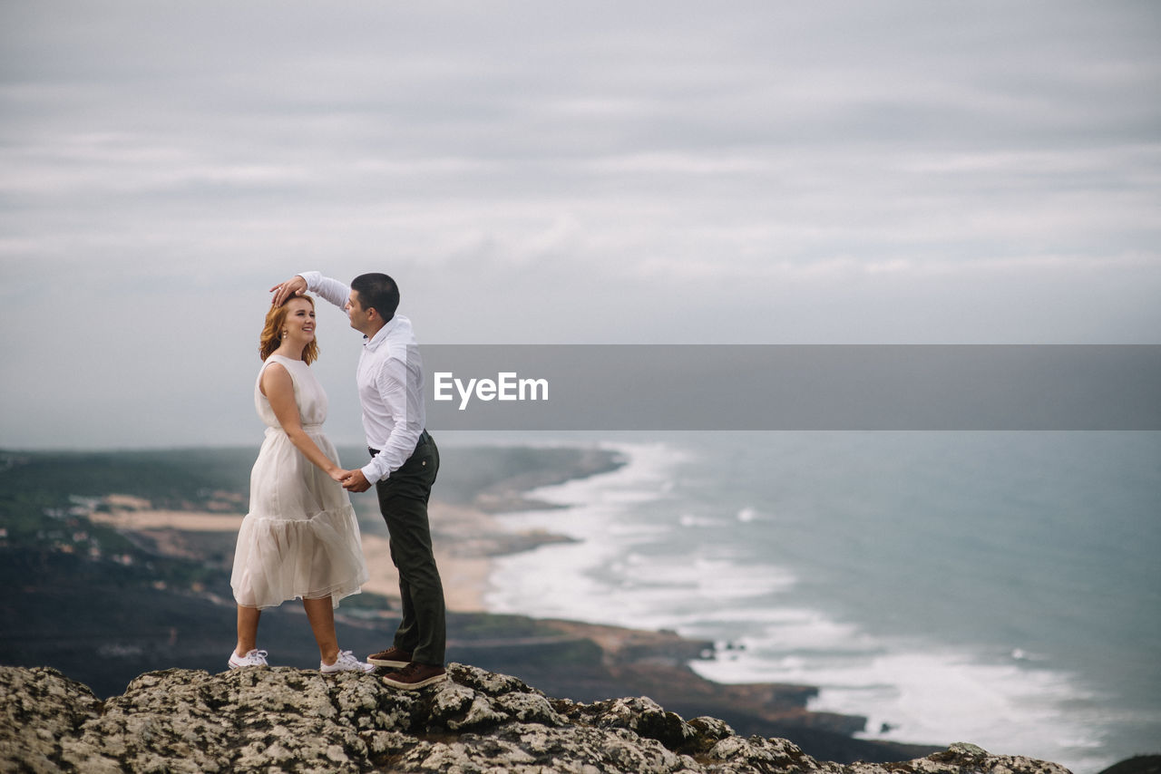 Romantic couple standing on cliff by sea against sky