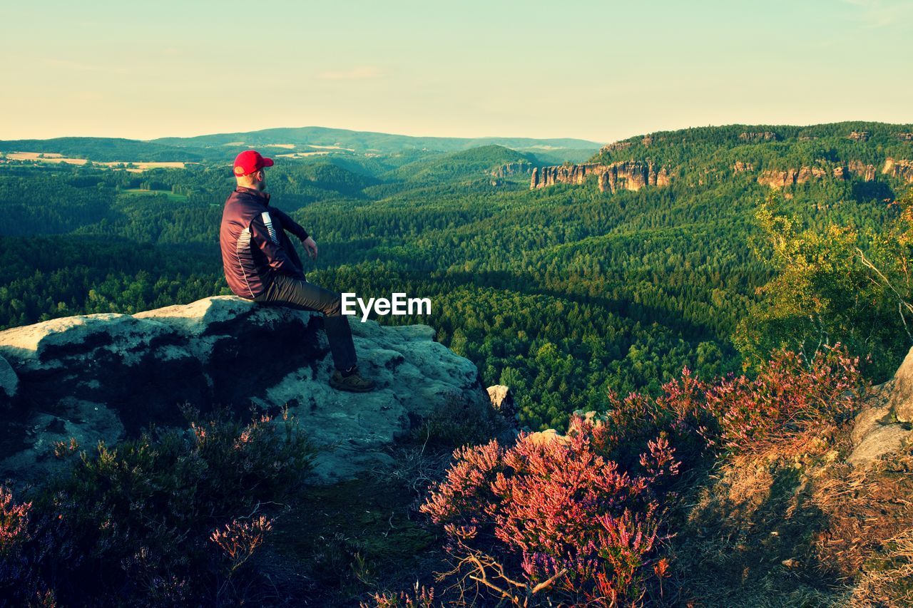 Tall man in black with red baseball cap on sharp cliff and watch to valley. morning in park.