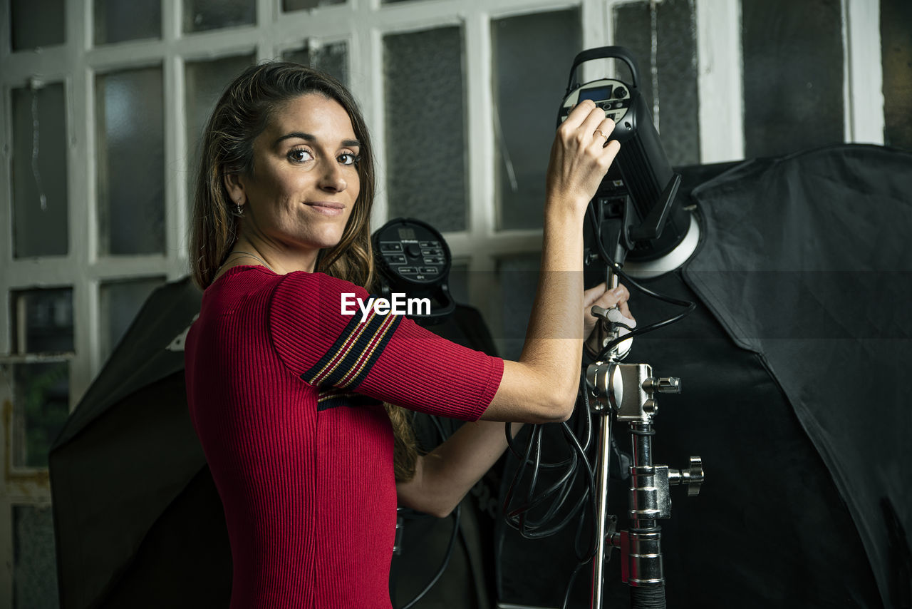 Portrait of smiling woman holding camera while standing in studio