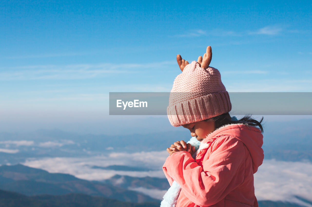 Girl praying while standing against sky during winter