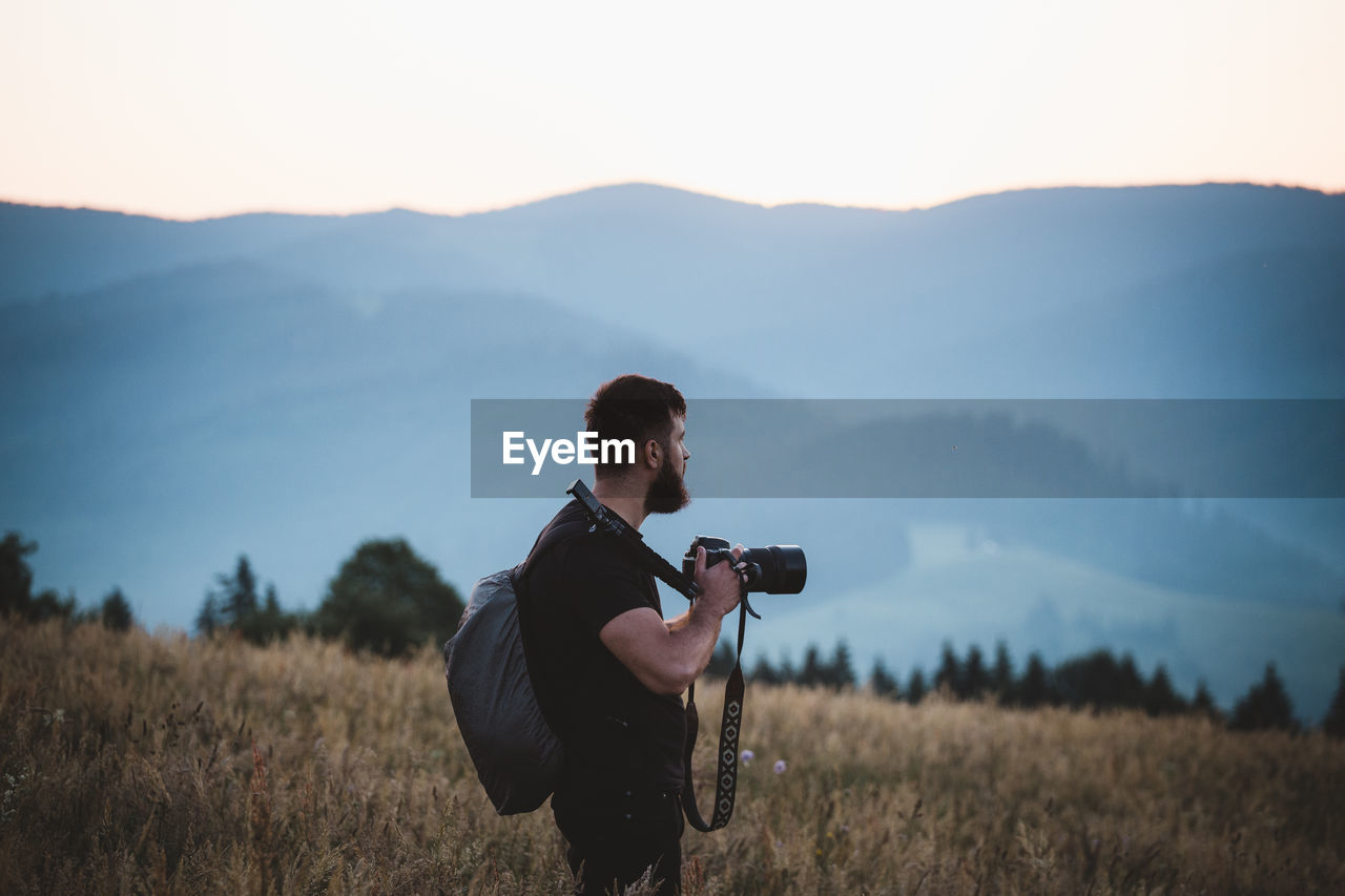 MAN PHOTOGRAPHING ON FIELD AGAINST MOUNTAIN