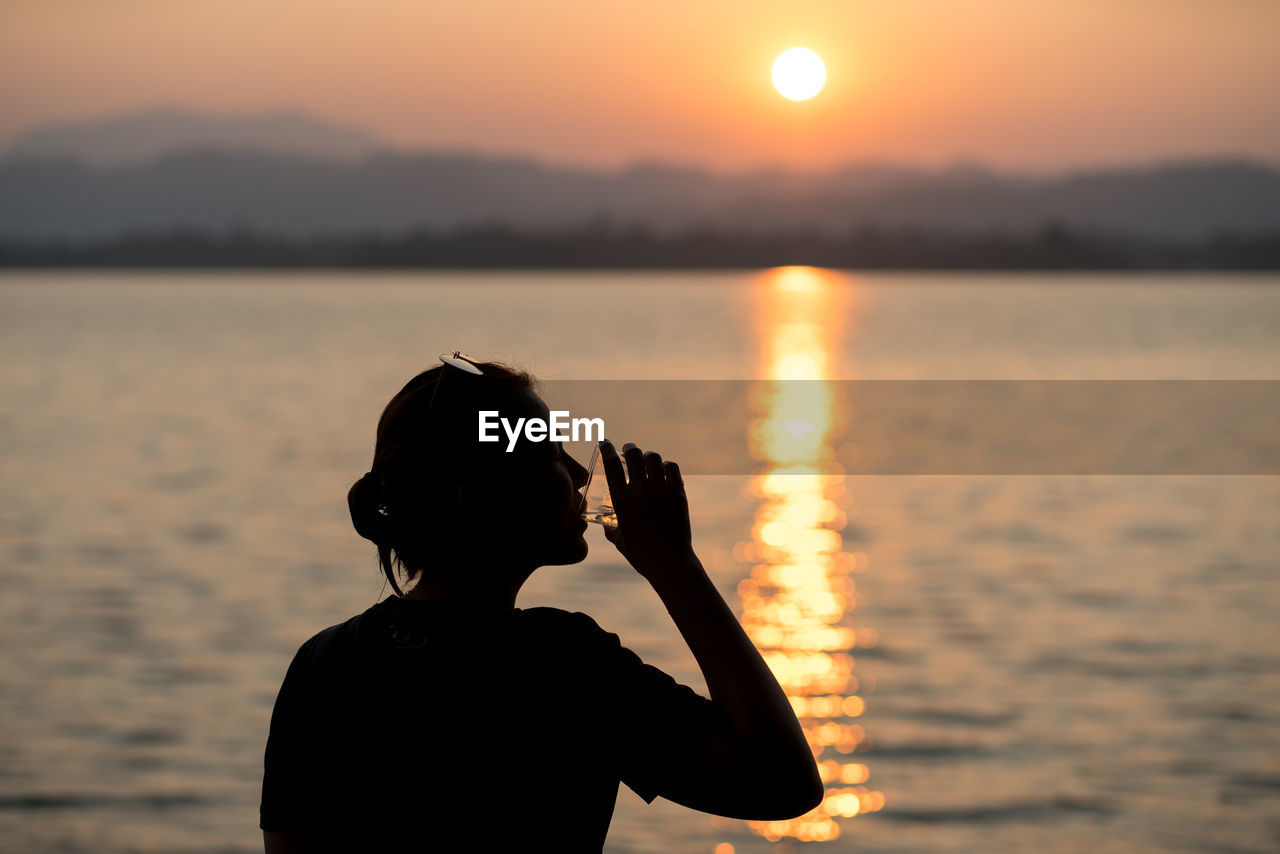 Silhouette woman drinking alcohol from glass while standing by sea against sky during sunset