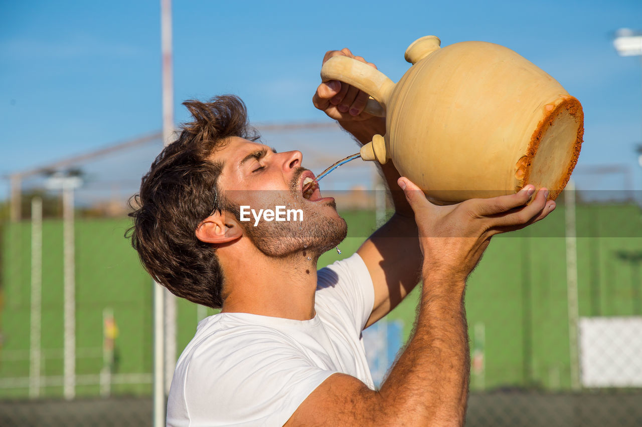 Young man drinking water from pot at court during sunny day