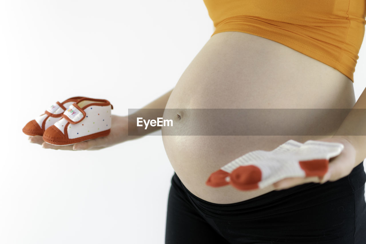 Midsection of pregnant woman holding baby booties and socks against white background