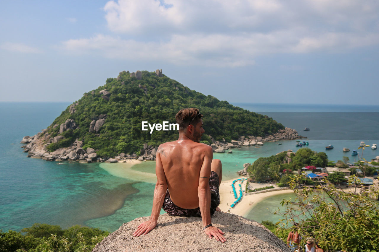 Rear view of shirtless young man looking at sea while sitting against sky