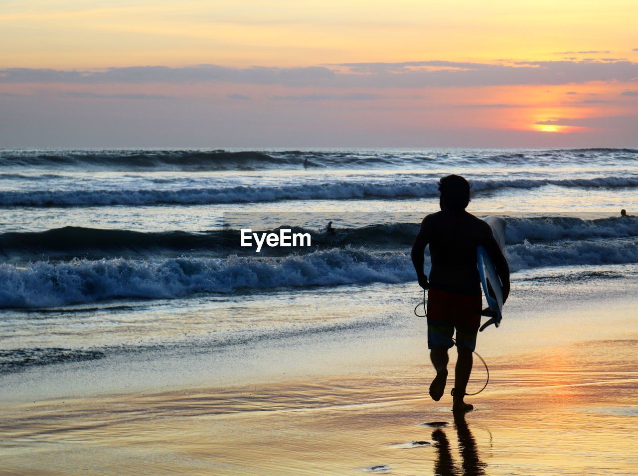 Rear view of man with surfboard running at beach during sunset