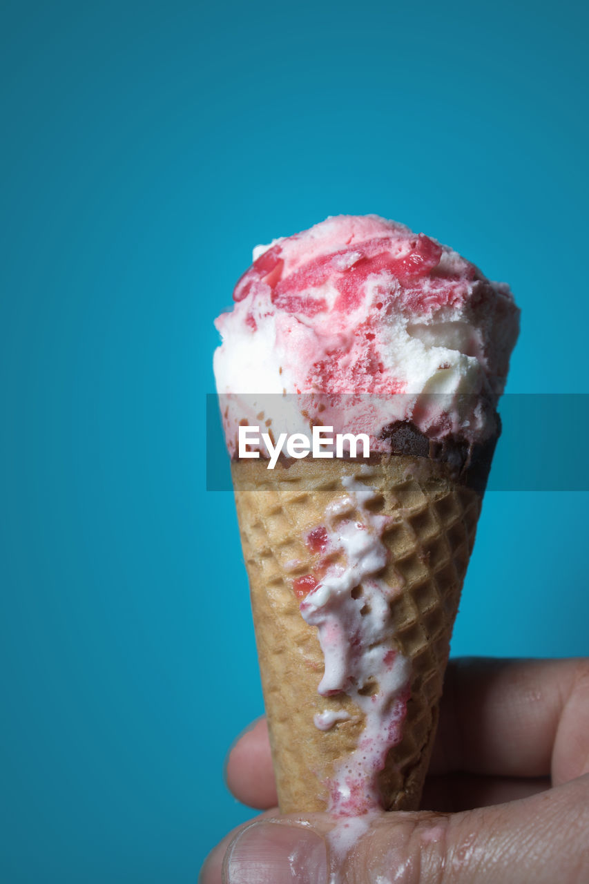 Cropped hand of person holding melting ice cream against blue background