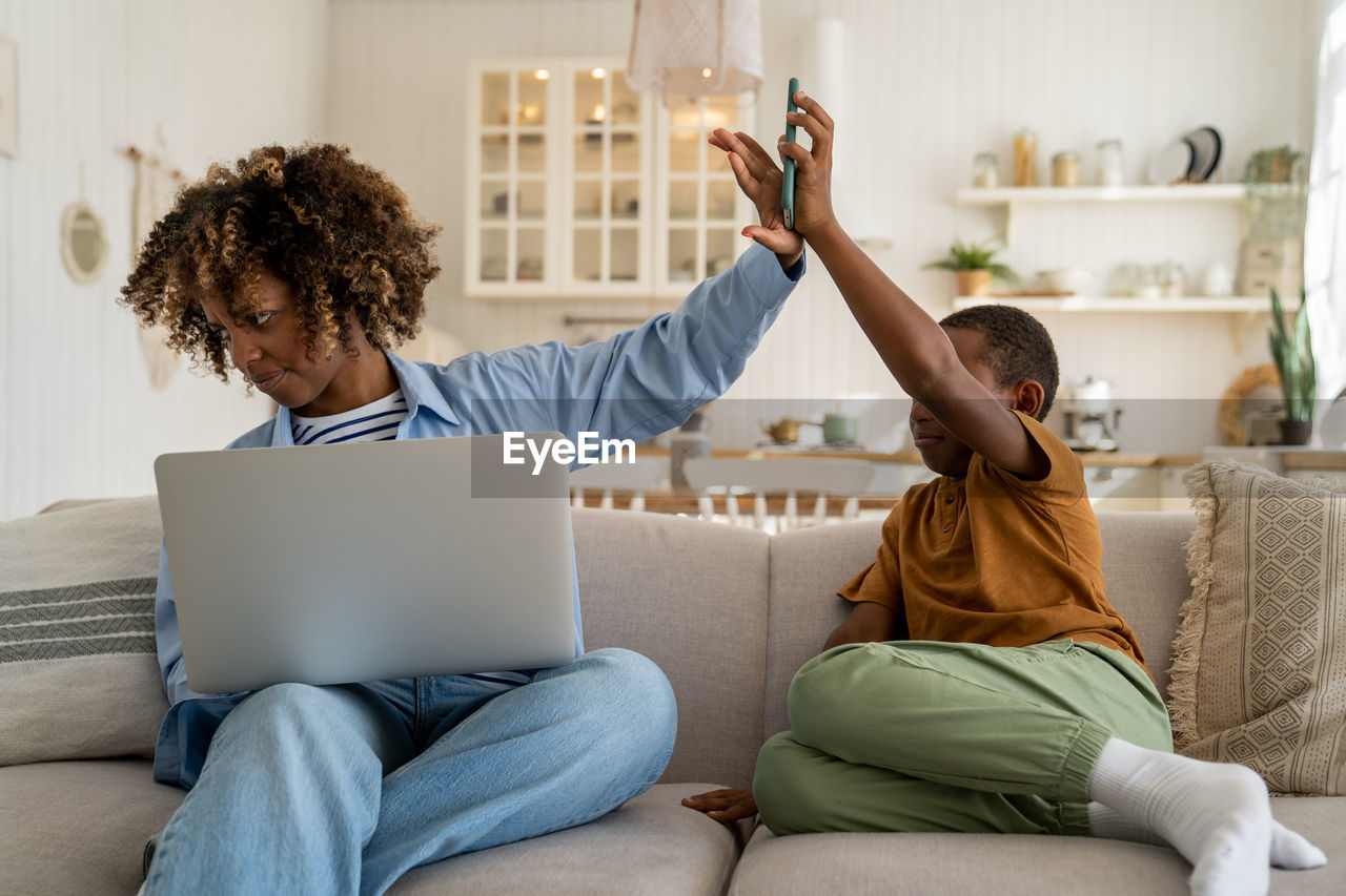 Nervous african american mother ignoring playful little boy son while working remotely on laptop