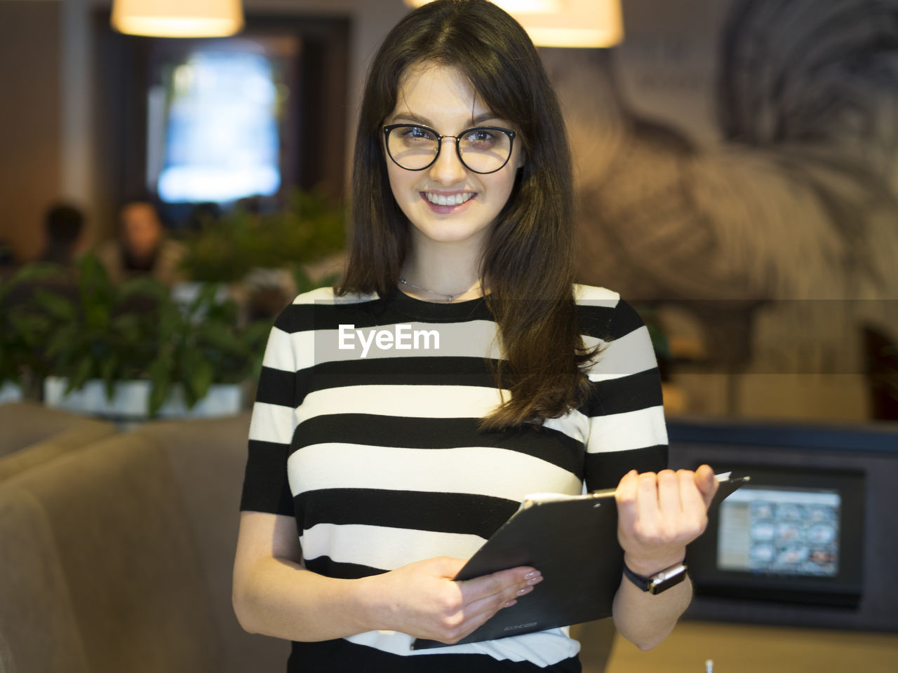 Portrait of confident young woman with clipboard standing in restaurant