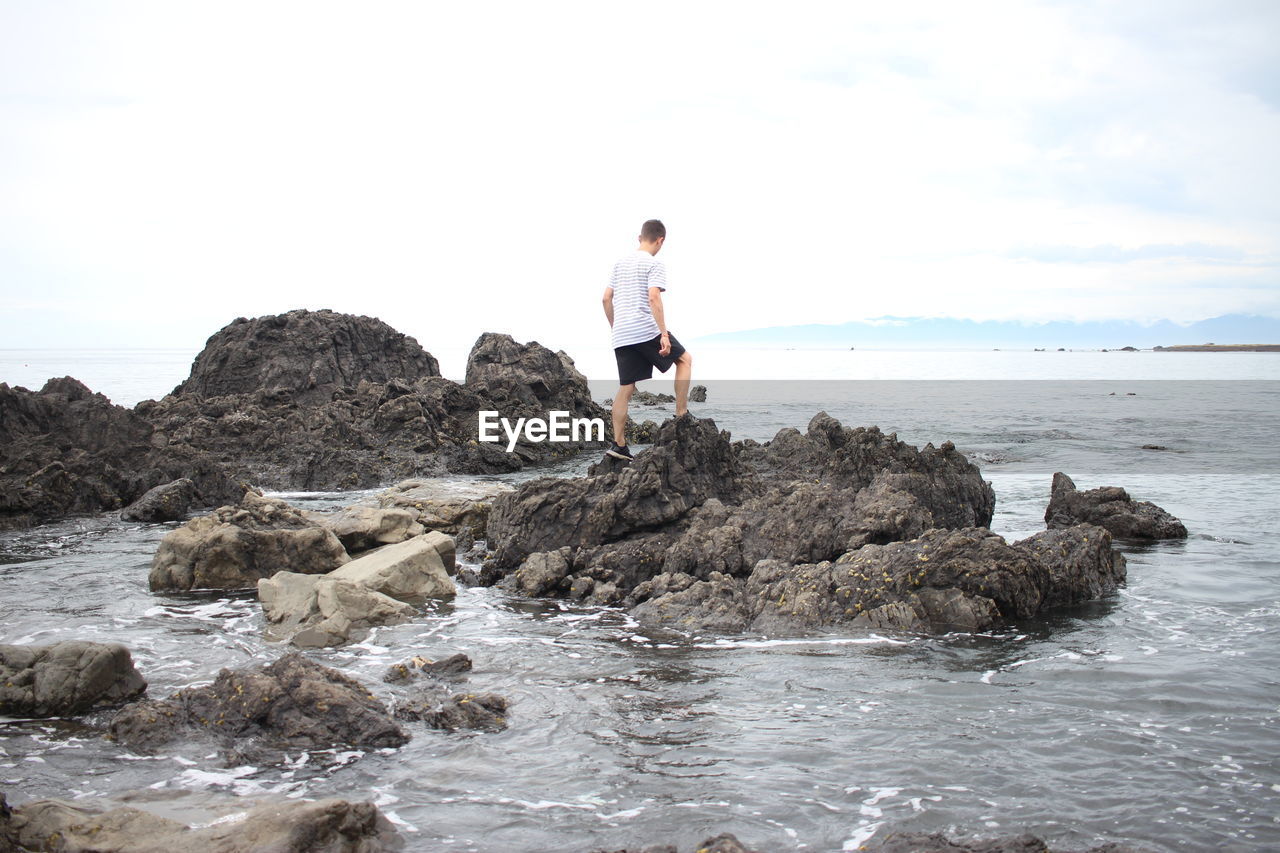 MAN ON ROCK AT BEACH AGAINST SKY