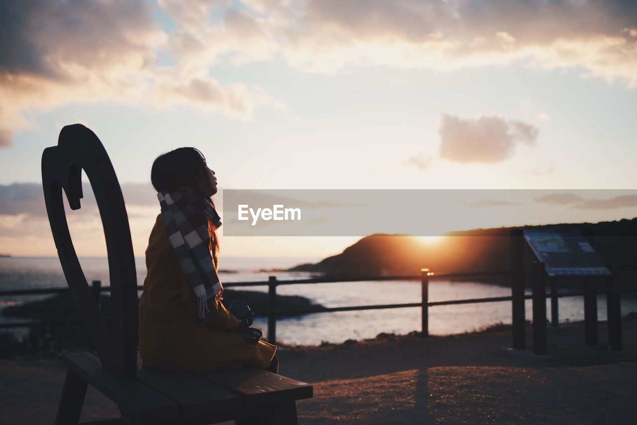 SIDE VIEW OF WOMAN SITTING BY SEA AGAINST SUNSET SKY