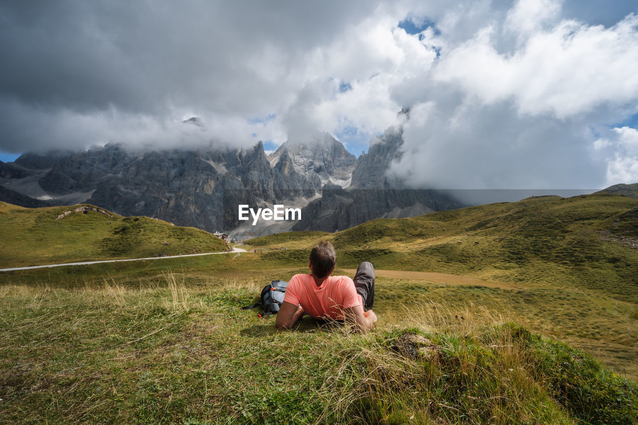 REAR VIEW OF MAN SITTING ON GRASS AGAINST MOUNTAIN RANGE
